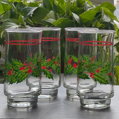 Set of four clear glasses with holly design on a reflective surface with green plants in the background.