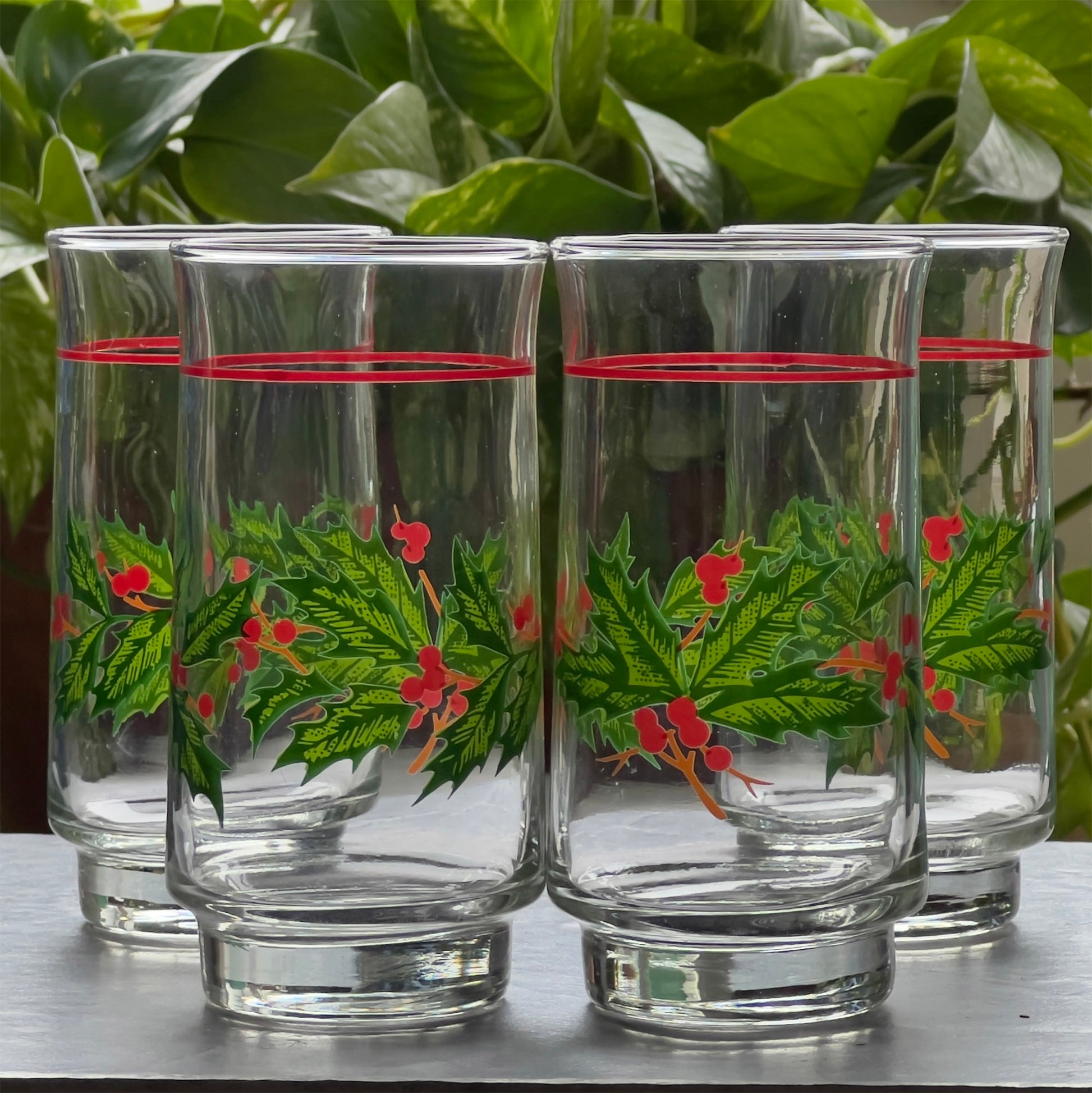 Four glass tumblers with holly leaf design on a wooden surface with green foliage in the background.