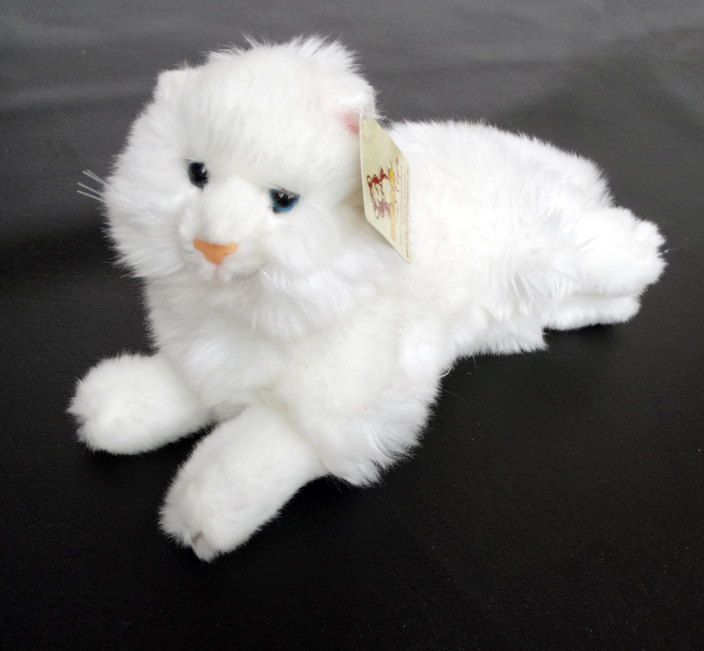 A white Angora plush cat toy with blue eyes, made of laying on a black surface.