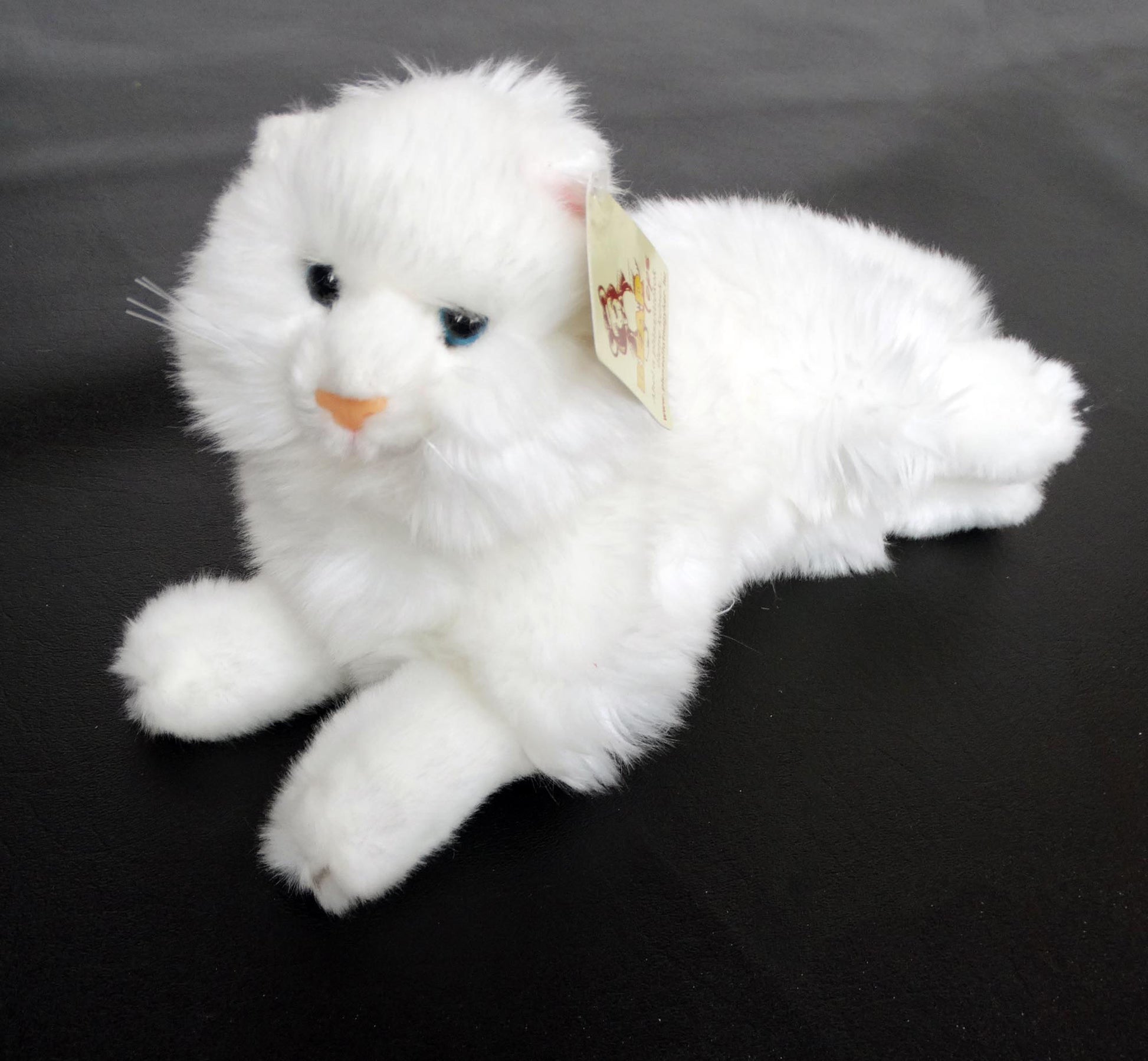 A white Angora plush cat toy with blue eyes, made of laying on a black surface.